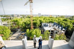 Two men wearing hard hats and holding building plans staning atop a building under construction